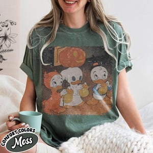 May include: Green t-shirt with a vintage Halloween graphic of three cartoon ducks in costumes, with a pumpkin. The shirt has rolled-up sleeves and the text "COMFORT COLORS Moss" on the bottom left.