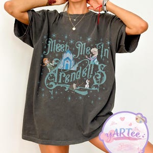 Pode incluir: T-shirt cinzento escuro com o texto "Meet Me In Arendelle" em turquesa. O design gr&aacute;fico inclui ilustra&ccedil;&otilde;es de personagens e um castelo do filme. A t-shirt tem mangas curtas e um corte folgado.