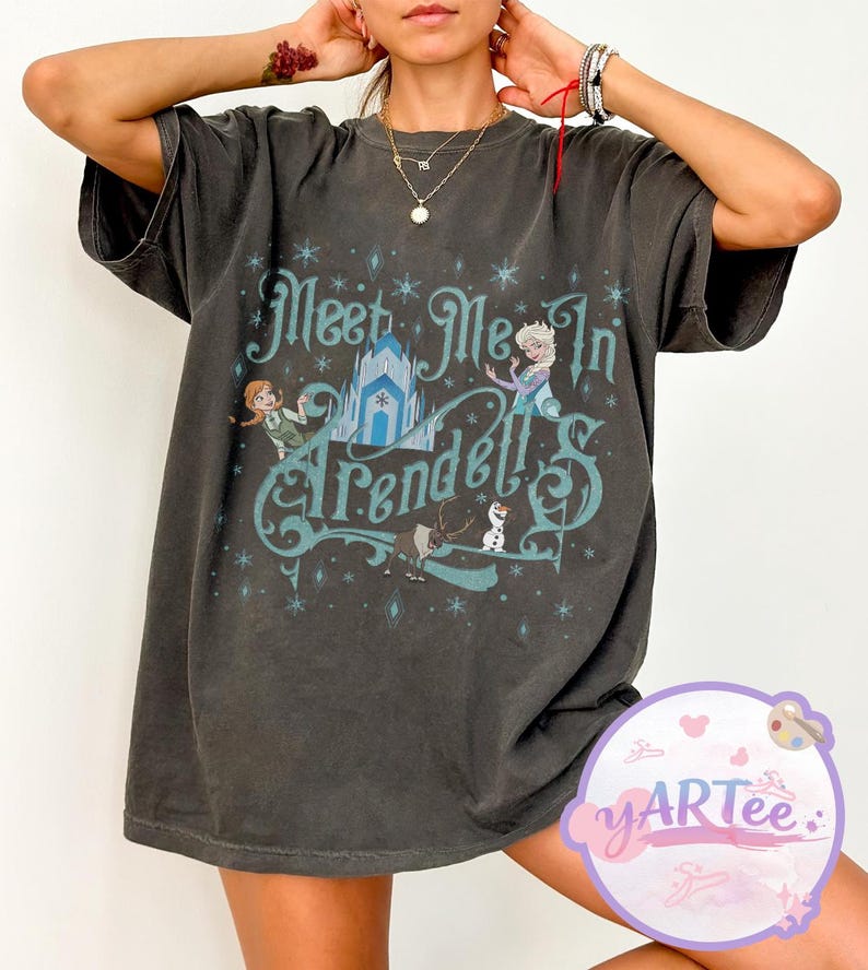 Pode incluir: T-shirt cinzento escuro com o texto "Meet Me In Arendelle" em turquesa. O design gr&aacute;fico inclui ilustra&ccedil;&otilde;es de personagens e um castelo do filme. A t-shirt tem mangas curtas e um corte folgado.