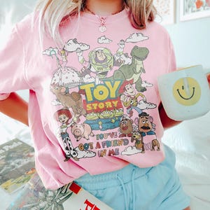 May include: Pink t-shirt featuring a colorful graphic of Toy Story characters, including Buzz Lightyear, Woody, and Rex. The text "Toy Story" and "You've Got a Friend in Me" are also visible. A white mug with a yellow smiley face is held in the hand.