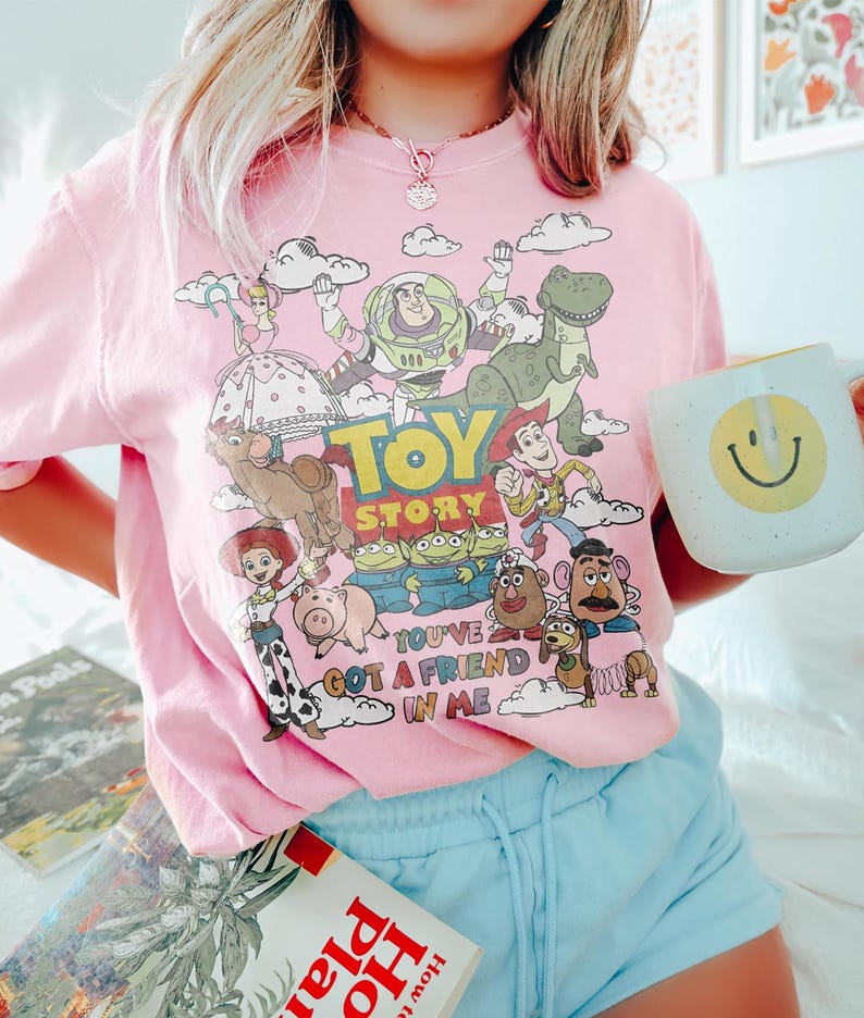 May include: Pink t-shirt featuring a colorful graphic of Toy Story characters, including Buzz Lightyear, Woody, and Rex. The text "Toy Story" and "You've Got a Friend in Me" are also visible. A white mug with a yellow smiley face is held in the hand.