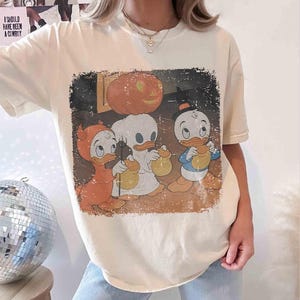 May include: Cream-coloured t-shirt with a vintage Halloween graphic. The graphic shows three cartoon ducks in costumes, a carved pumpkin, and the text "I SHOULD HAVE BEEN A COWBOY".
