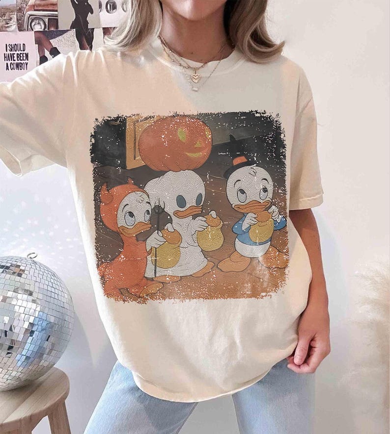 May include: Cream-coloured t-shirt with a vintage Halloween graphic. The graphic shows three cartoon ducks in costumes, a carved pumpkin, and the text "I SHOULD HAVE BEEN A COWBOY".