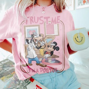 May include: Pink t-shirt featuring a cartoon illustration of Goofy and Minnie Mouse in a doctor's office. The text "TRUST ME I'M A DOCTOR" is printed above and below the graphic. A white mug with a yellow smiley face is held in the hand.