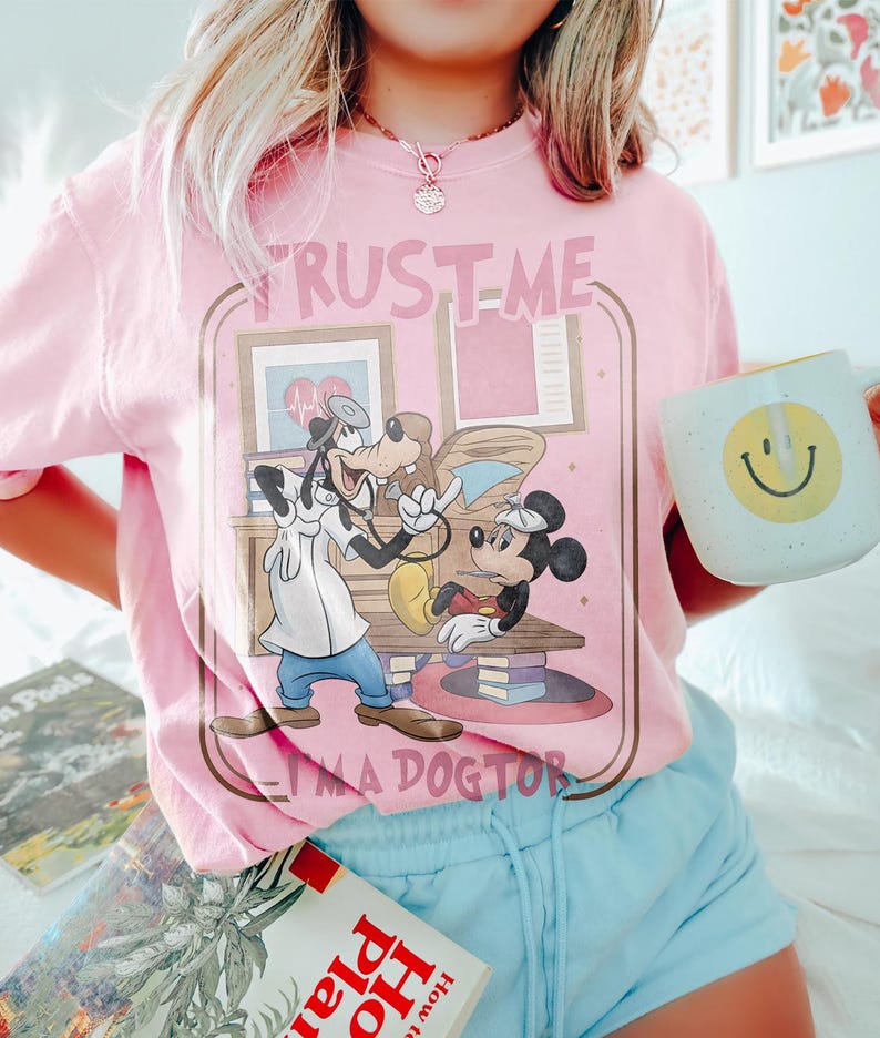 May include: Pink t-shirt featuring a cartoon illustration of Goofy and Minnie Mouse in a doctor's office. The text "TRUST ME I'M A DOCTOR" is printed above and below the graphic. A white mug with a yellow smiley face is held in the hand.
