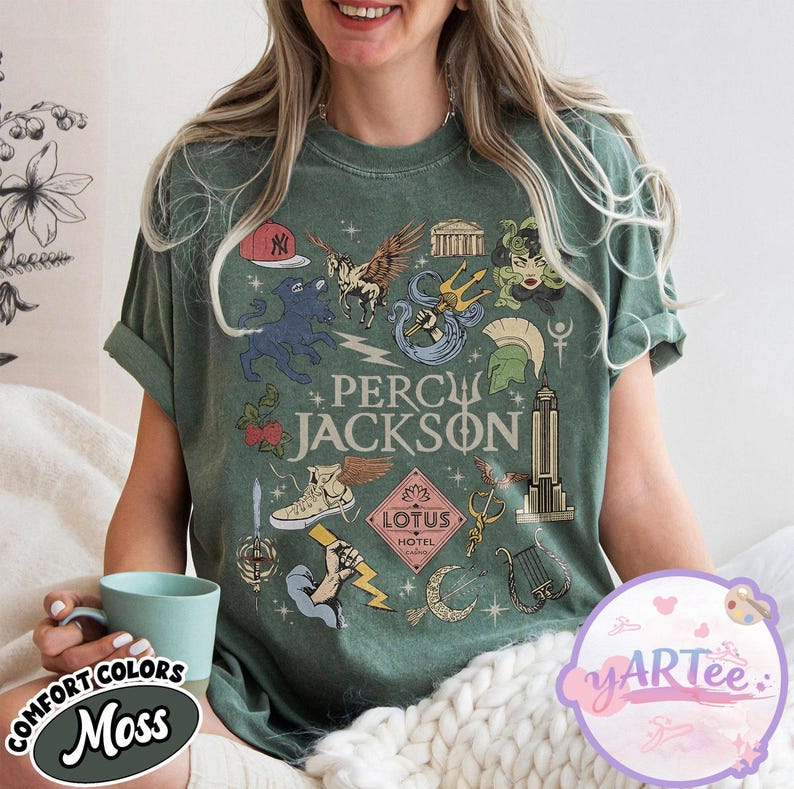Pu&ograve; includere: T-shirt verde muschio con un design grafico con la scritta "PERCY JACKSON" e varie illustrazioni. Il design include un berretto da baseball, Pegaso, un tridente, Medusa e l'Empire State Building.