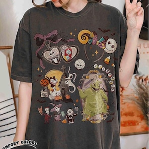 May include: Dark gray t-shirt featuring a colorful graphic design with characters and elements from "The Nightmare Before Christmas." The design includes Jack Skellington, Sally, Oogie Boogie, and other Halloween-themed imagery. The shirt has the text "Nightmare" and "Comfort Colors Pepper".