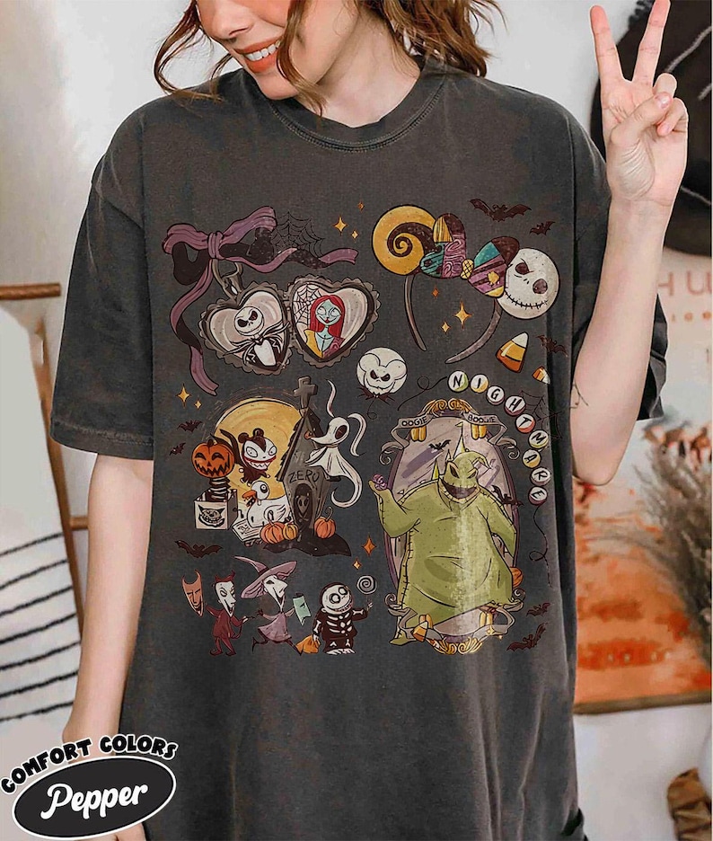 May include: Dark gray t-shirt featuring a colorful graphic design with characters and elements from "The Nightmare Before Christmas." The design includes Jack Skellington, Sally, Oogie Boogie, and other Halloween-themed imagery. The shirt has the text "Nightmare" and "Comfort Colors Pepper".