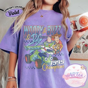 May include: Violet-colored t-shirt featuring a graphic of Woody, Buzz Lightyear, and RC from Toy Story. The design includes the text "WOODY & BUZZ & RC" and "1995 Champions". The shirt has a vintage, distressed look.