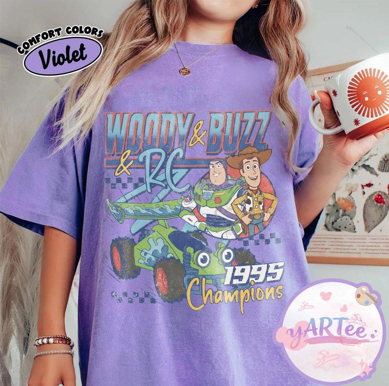 May include: Violet-colored t-shirt featuring a graphic of Woody, Buzz Lightyear, and RC from Toy Story. The design includes the text "WOODY & BUZZ & RC" and "1995 Champions". The shirt has a vintage, distressed look.