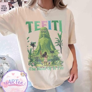 K&ouml;nnte beinhalten: Cremefarbenes T-Shirt mit einer Grafik von Te Fiti aus dem Film Vaiana. Das Design zeigt das Wort "TE FITI" in Gr&uuml;n und Rosa, mit dem Satz "The Head Of Te Fiti" unter dem Bild der Figur.