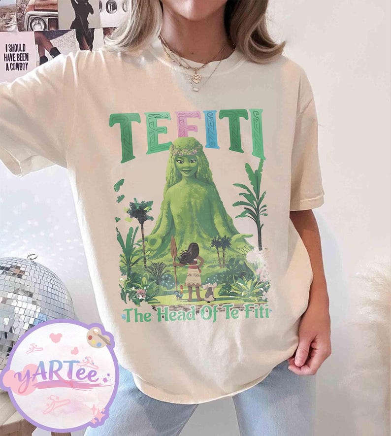 K&ouml;nnte beinhalten: Cremefarbenes T-Shirt mit einer Grafik von Te Fiti aus dem Film Vaiana. Das Design zeigt das Wort "TE FITI" in Gr&uuml;n und Rosa, mit dem Satz "The Head Of Te Fiti" unter dem Bild der Figur.
