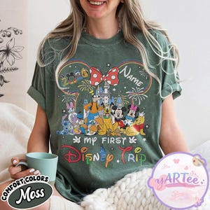 May include: Green t-shirt with a colorful Disney graphic. The design includes a castle, fireworks, and various Disney characters. Text on the shirt reads "My First Disney Trip" and "Name". The shirt has a round neck and short sleeves.