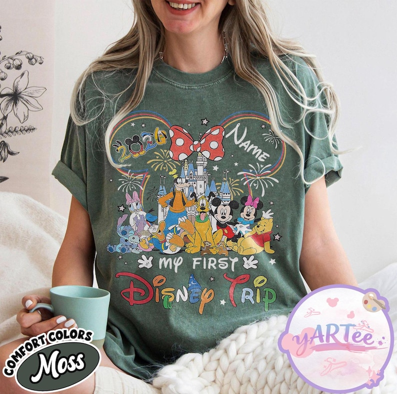 May include: Green t-shirt with a colorful Disney graphic. The design includes a castle, fireworks, and various Disney characters. Text on the shirt reads "My First Disney Trip" and "Name". The shirt has a round neck and short sleeves.