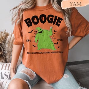 May include: Orange t-shirt featuring the word "BOOGIE" above a green Oogie Boogie character from "The Nightmare Before Christmas." The design includes bats, a spiderweb, and the text "EST. 1993."