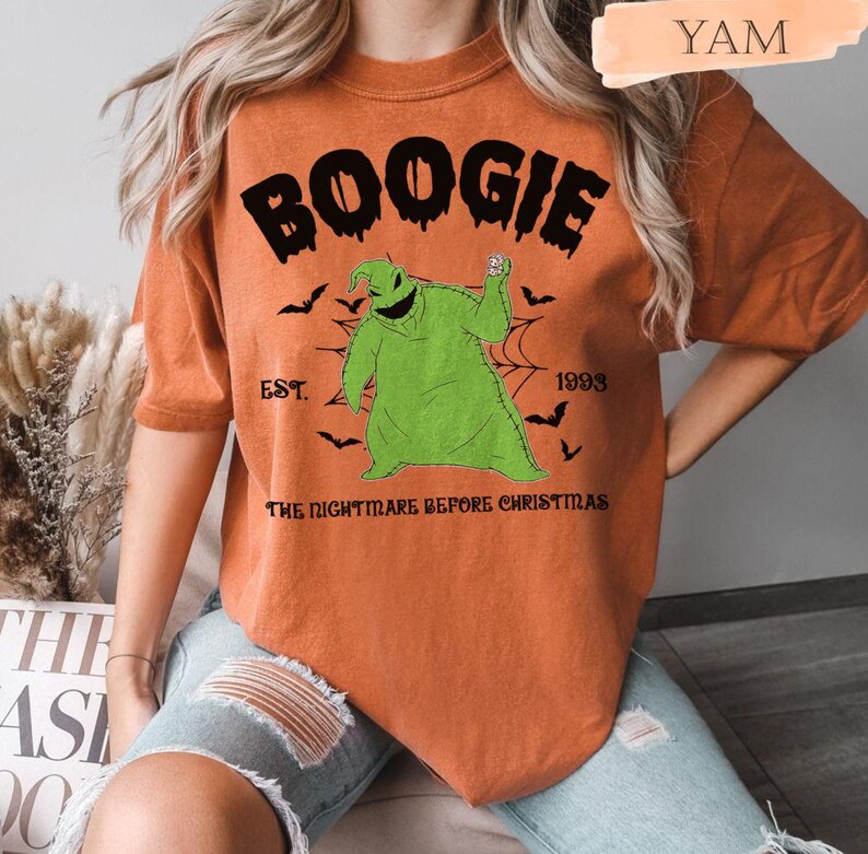 May include: Orange t-shirt featuring the word "BOOGIE" above a green Oogie Boogie character from "The Nightmare Before Christmas." The design includes bats, a spiderweb, and the text "EST. 1993."
