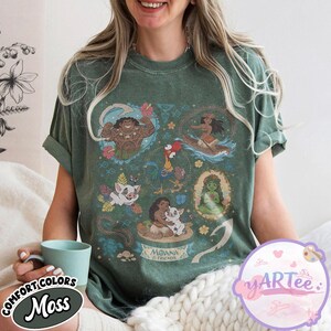 K&ouml;nnte beinhalten: Moosgr&uuml;nes T-Shirt mit einer farbenfrohen Grafik von Charakteren aus dem Animationsfilm Moana. Das Design enth&auml;lt Bilder von Maui, Moana, Pua, Hei Hei und anderen Charakteren. Das Shirt tr&auml;gt die Aufschrift "Moana & Friends".