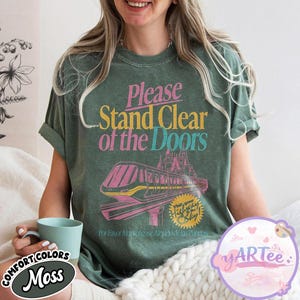 May include: A moss green t-shirt with the text "Please Stand Clear of the Doors" in pink and yellow. The shirt features a pink and yellow monorail graphic and the words "Visit your Highway in the Sky!"