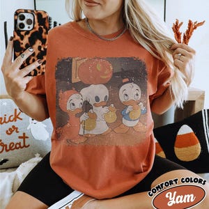 May include: A rust-coloured t-shirt with a vintage Halloween graphic of three cartoon ducks in costumes, holding treat bags. The shirt has a distressed look, with the words "Comfort Colors" and "Yam" in a logo on the bottom right.