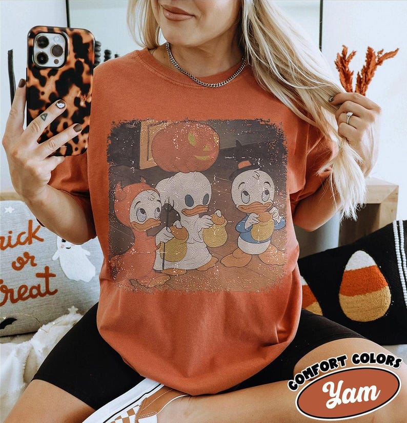 May include: A rust-coloured t-shirt with a vintage Halloween graphic of three cartoon ducks in costumes, holding treat bags. The shirt has a distressed look, with the words "Comfort Colors" and "Yam" in a logo on the bottom right.