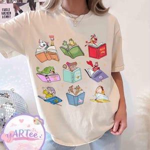 Puede incluir: Camiseta color crema con un gr&aacute;fico colorido de personajes de dibujos animados leyendo libros. El dise&ntilde;o incluye personajes como Moana y Rapunzel, cada uno con su propio libro. El texto "I SHOULD HAVE BEEN A COWBOY" es visible en el fondo.