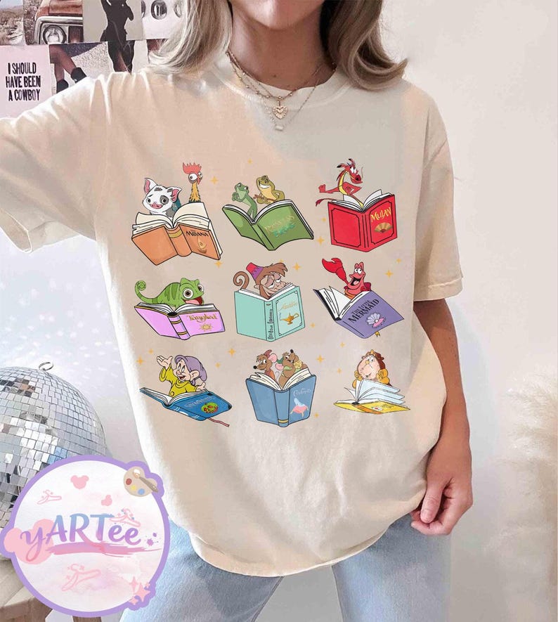 Puede incluir: Camiseta color crema con un gr&aacute;fico colorido de personajes de dibujos animados leyendo libros. El dise&ntilde;o incluye personajes como Moana y Rapunzel, cada uno con su propio libro. El texto "I SHOULD HAVE BEEN A COWBOY" es visible en el fondo.
