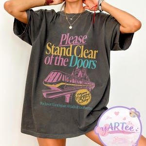 May include: Dark gray t-shirt with the text "Please Stand Clear of the Doors" in pink, yellow, and teal. A graphic of a monorail and castle is below the text. The shirt also has text in Spanish.
