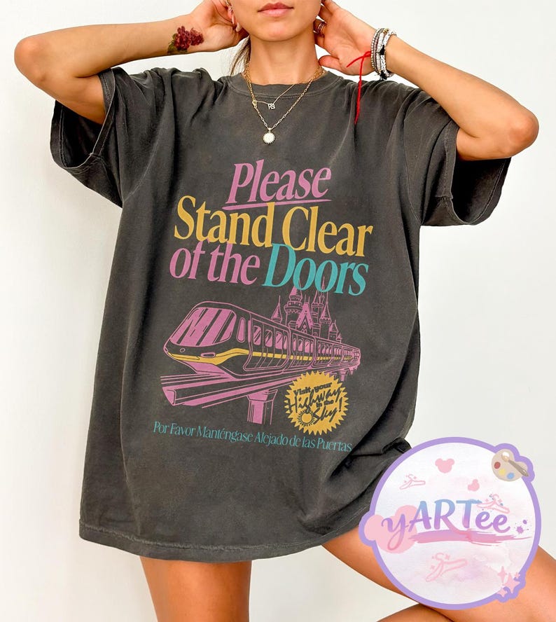 May include: Dark gray t-shirt with the text "Please Stand Clear of the Doors" in pink, yellow, and teal. A graphic of a monorail and castle is below the text. The shirt also has text in Spanish.