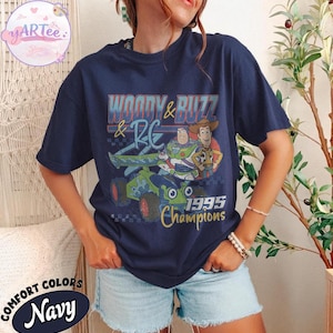 May include: Navy blue t-shirt featuring a graphic of Woody and Buzz Lightyear from Toy Story, with the text "Woody & Buzz & RC 1995 Champions." The shirt is a Comfort Colors brand.