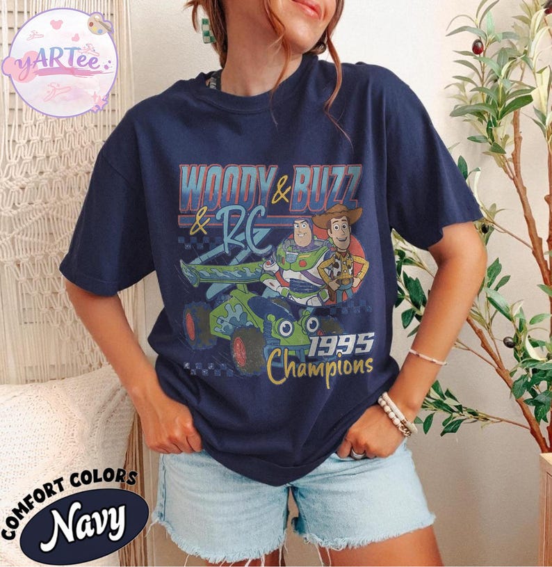 May include: Navy blue t-shirt featuring a graphic of Woody and Buzz Lightyear from Toy Story, with the text "Woody & Buzz & RC 1995 Champions." The shirt is a Comfort Colors brand.