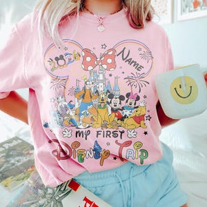 May include: Pink t-shirt with a Disney theme. The shirt features a graphic of Disney characters, the text "My First Disney Trip", and the year 2020. A white mug with a yellow smiley face is held in the hand.