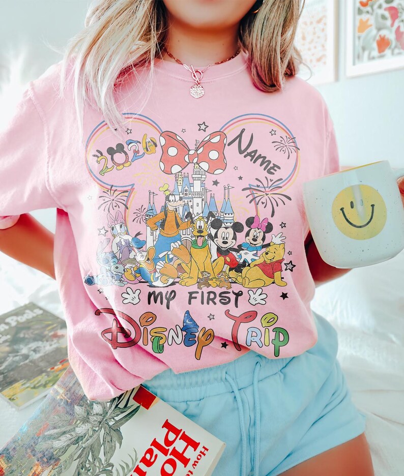May include: Pink t-shirt with a Disney theme. The shirt features a graphic of Disney characters, the text "My First Disney Trip", and the year 2020. A white mug with a yellow smiley face is held in the hand.