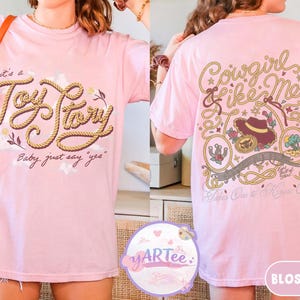 May include: A light pink t-shirt featuring a front graphic with the text "it's a Toy Story Baby, just say "yes"" in rope-style lettering. The back graphic displays "Cowgirl like Me" with a cowboy hat, lasso, and "Takes One to Know One" text.