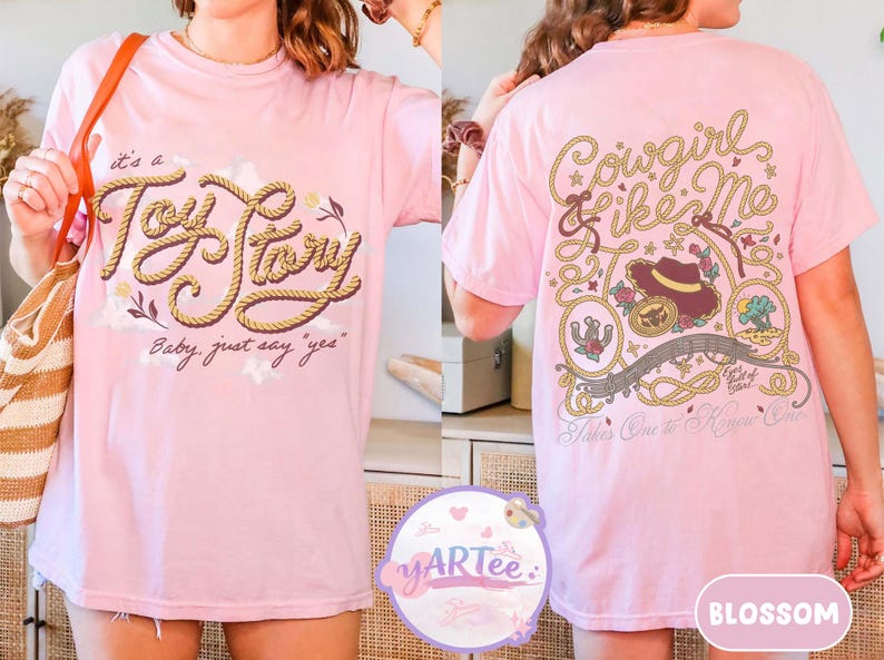 May include: A light pink t-shirt featuring a front graphic with the text "it's a Toy Story Baby, just say "yes"" in rope-style lettering. The back graphic displays "Cowgirl like Me" with a cowboy hat, lasso, and "Takes One to Know One" text.