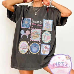 May include: Dark gray t-shirt featuring a colorful graphic design with the text "Meet Me On Main Street." The design includes various themed illustrations, such as a castle, a ferris wheel, and a rose. The shirt has short sleeves and a relaxed fit.