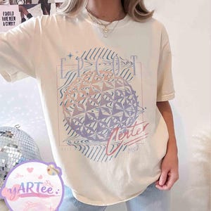 May include: Cream-colored t-shirt featuring a graphic of a geometric sphere with the text "EPCOT Center" and "Winter 1982". The design incorporates shades of pink, purple, and blue. The shirt has short sleeves.