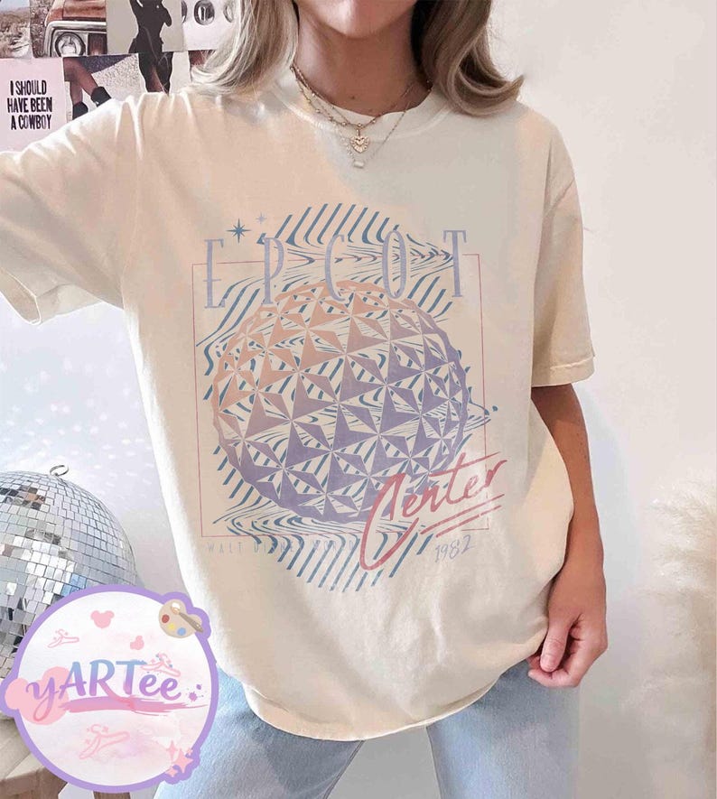 May include: Cream-colored t-shirt featuring a graphic of a geometric sphere with the text "EPCOT Center" and "Winter 1982". The design incorporates shades of pink, purple, and blue. The shirt has short sleeves.