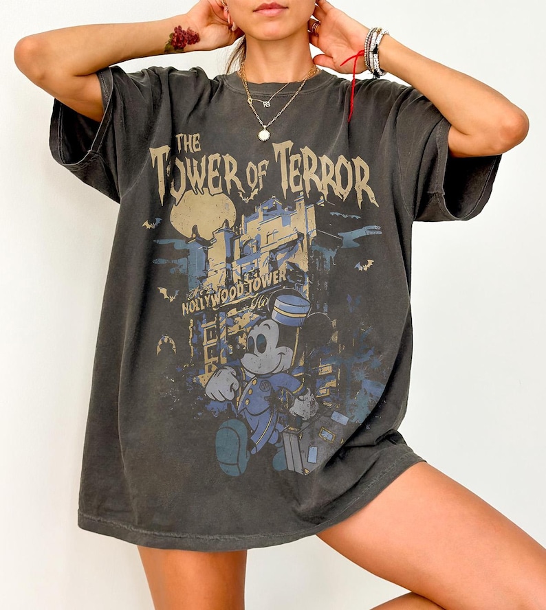 May include: Dark gray t-shirt featuring a graphic of Mickey Mouse in a bellhop uniform in front of the Hollywood Tower Hotel. The text "The Tower of Terror" and "Hollywood Tower Hotel" are printed above and below the image.