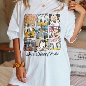 Op de afbeelding: Wit overformaat T-shirt met een grafische print van verschillende Disney-figuren, waaronder Mickey Mouse, Minnie Mouse, Donald Duck, Goofy en Pluto. Het shirt heeft ook de tekst "Walt Disney World" en "Hollywood & Vine".