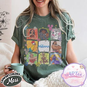 May include: Moss green t-shirt featuring a colorful graphic of characters from the Disney movie Moana. The design includes images of Moana, Maui, Hei Hei, and Pua. The shirt also has the words "Disney Moana" printed on it.
