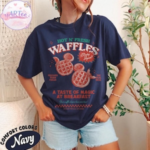 Puede incluir: Camiseta azul marino con un gr&aacute;fico que presenta el texto "HOT N' FRESH WAFFLES" y dos gofres con forma de Mickey Mouse. La camiseta tambi&eacute;n incluye las frases "Fresher Than Fresh" y "A Taste of Magic at Breakfast".