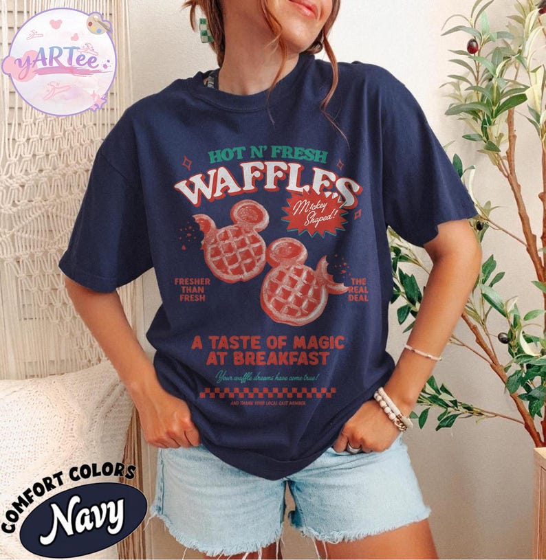 Puede incluir: Camiseta azul marino con un gr&aacute;fico que presenta el texto "HOT N' FRESH WAFFLES" y dos gofres con forma de Mickey Mouse. La camiseta tambi&eacute;n incluye las frases "Fresher Than Fresh" y "A Taste of Magic at Breakfast".