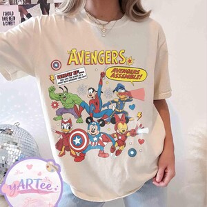 May include: Cream-colored t-shirt featuring a colorful graphic of The Avengers characters, including Hulk, Spider-Man, and Mickey Mouse, in a comic book style. Text includes "AVENGERS" and "AVENGERS ASSEMBLE!"