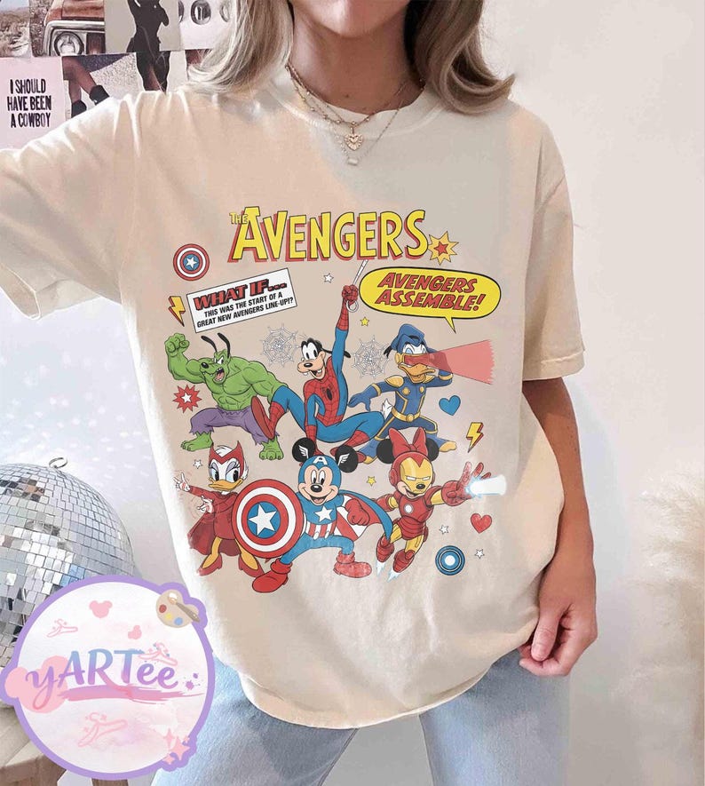 May include: Cream-colored t-shirt featuring a colorful graphic of The Avengers characters, including Hulk, Spider-Man, and Mickey Mouse, in a comic book style. Text includes "AVENGERS" and "AVENGERS ASSEMBLE!"