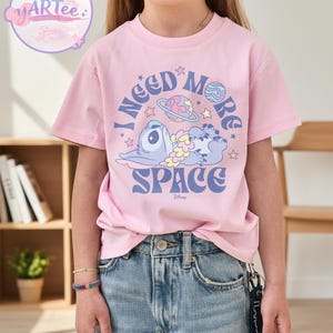 May include: Pink t-shirt featuring a cartoon character and the text "I NEED MORE SPACE" in blue. The design includes planets and stars. The shirt is worn by a child.
