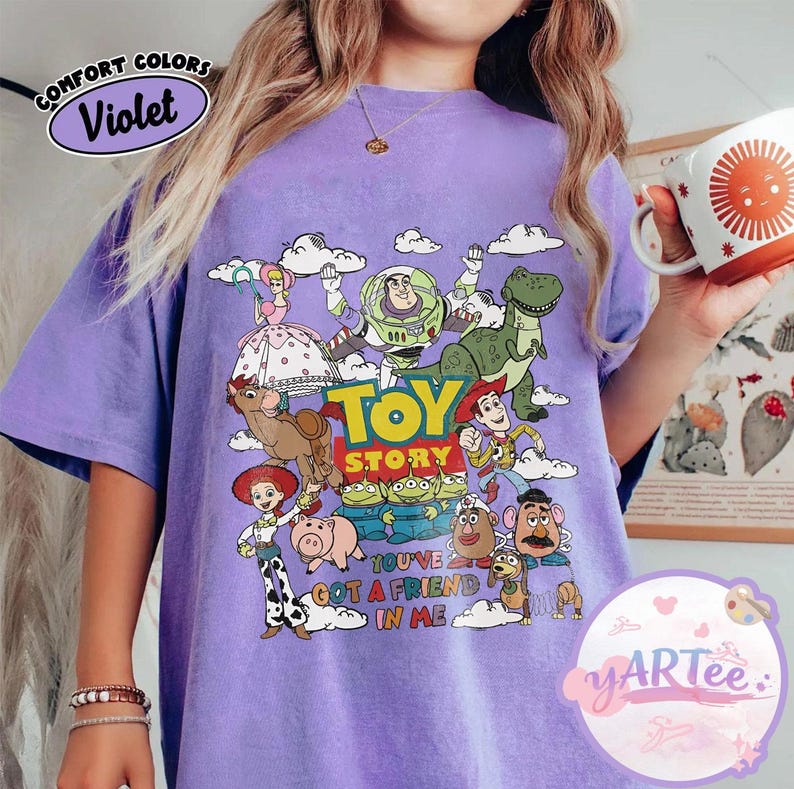 May include: Violet-colored t-shirt featuring a graphic of characters from the animated film "Toy Story." The design includes Woody, Buzz Lightyear, Rex, and other characters, with the text "TOY STORY" and "YOU'VE GOT A FRIEND IN ME."