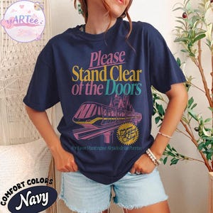 May include: Navy blue t-shirt with the text "Please Stand Clear of the Doors" in pink, yellow, and teal. A graphic of a monorail and castle is below the text. The shirt also has the text "Highway to Sly" and "For Favor Mant&eacute;ngase Alejado de las Puertas".