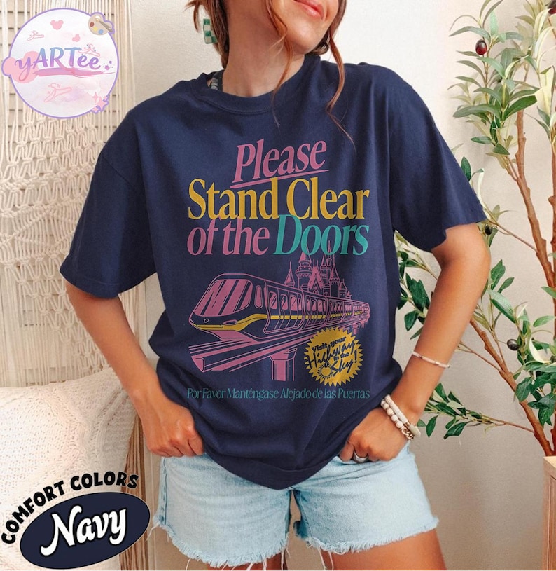 May include: Navy blue t-shirt with the text "Please Stand Clear of the Doors" in pink, yellow, and teal. A graphic of a monorail and castle is below the text. The shirt also has the text "Highway to Sly" and "For Favor Mant&eacute;ngase Alejado de las Puertas".
