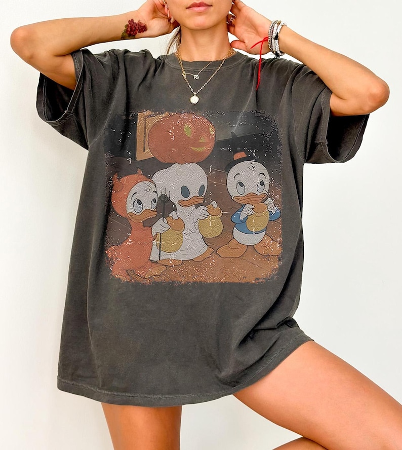 May include: Dark grey t-shirt with a Halloween graphic. The design shows three cartoon ducks in costumes, a pumpkin, and a spooky backdrop. The tee has a vintage, worn appearance.