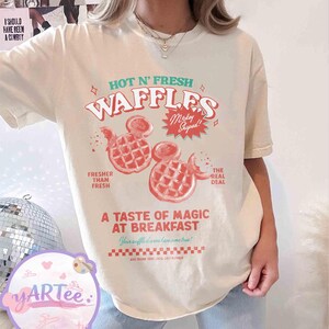 Puede incluir: Camiseta color crema con un dise&ntilde;o gr&aacute;fico. El dise&ntilde;o presenta el texto "HOT N' FRESH WAFFLES" y dos gofres con forma de Mickey Mouse en rojo. La camiseta tambi&eacute;n incluye las frases "Fresher Than Fresh" y "The Real Deal".